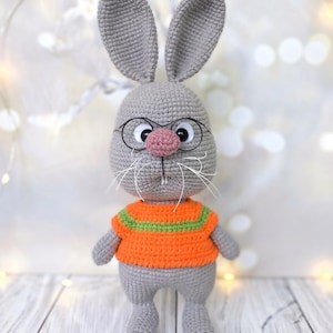 Puede incluir: Un conejo de ganchillo gris hecho a mano con gafas y un suéter naranja con rayas verdes. El conejo tiene una nariz rosa y bigotes blancos. El juguete está sobre una superficie clara.