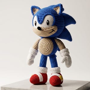 May include: A handcrafted crochet doll of a blue hedgehog character with large pointed ears, a tan muzzle, and a spiral pattern on the belly. The figure features white gloves, red shoes with white cuffs, and a black nose, standing upright on a flat surface.
