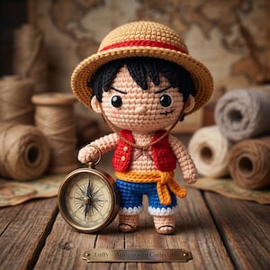 Op de afbeelding: Een gehaakte amigurumi-pop van een personage met een strooien hoed met rode band, een rood vest, een blauwe broek en een gele sjerp. De pop heeft zwart haar, een litteken onder het oog en houdt een koperen kompas vast. Een metalen plaatje vermeldt Luffy - Amigurumi Collectible.