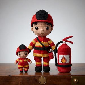 Pode incluir: Bonecos de bombeiro de crochê, um grande e um pequeno, ao lado de um extintor de incêndio vermelho de crochê. Os bonecos usam uniformes vermelhos e amarelos e capacetes pretos. O extintor tem um rótulo branco com uma chama.