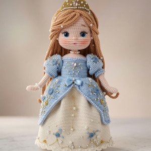 May include: A handcrafted crochet doll with long light brown hair, wearing a gold crown with pearls and a blue and cream gown. The dress features puff sleeves, blue floral embroidery, small white pearls, and a light blue bow at the waist.