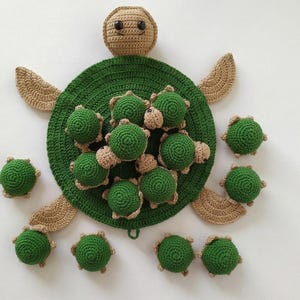 May include: A large crocheted turtle with a green shell and tan head and flippers, featuring a collection of ten smaller, matching crocheted baby turtles resting on top of the large shell and scattered around the perimeter of the main body.