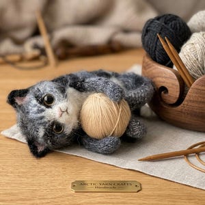 May include: A needle-felted gray and white kitten figurine holding a small beige yarn ball, positioned next to a wooden yarn bowl filled with various yarn skeins, circular knitting needles, and a brass plaque labeled ARCTIC YARN CRAFTS Handmade.