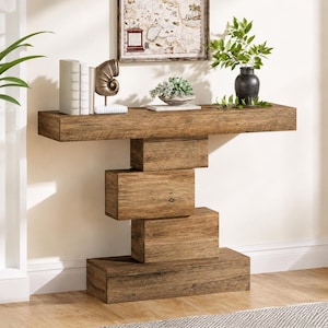 Peut inclure: Une console en bois rustique avec un design géométrique unique. La table présente un long plateau rectangulaire soutenu par des éléments empilés en forme de boîte. Des objets décoratifs tels que des livres, une sculpture de coquillage et un vase avec de la verdure sont exposés.