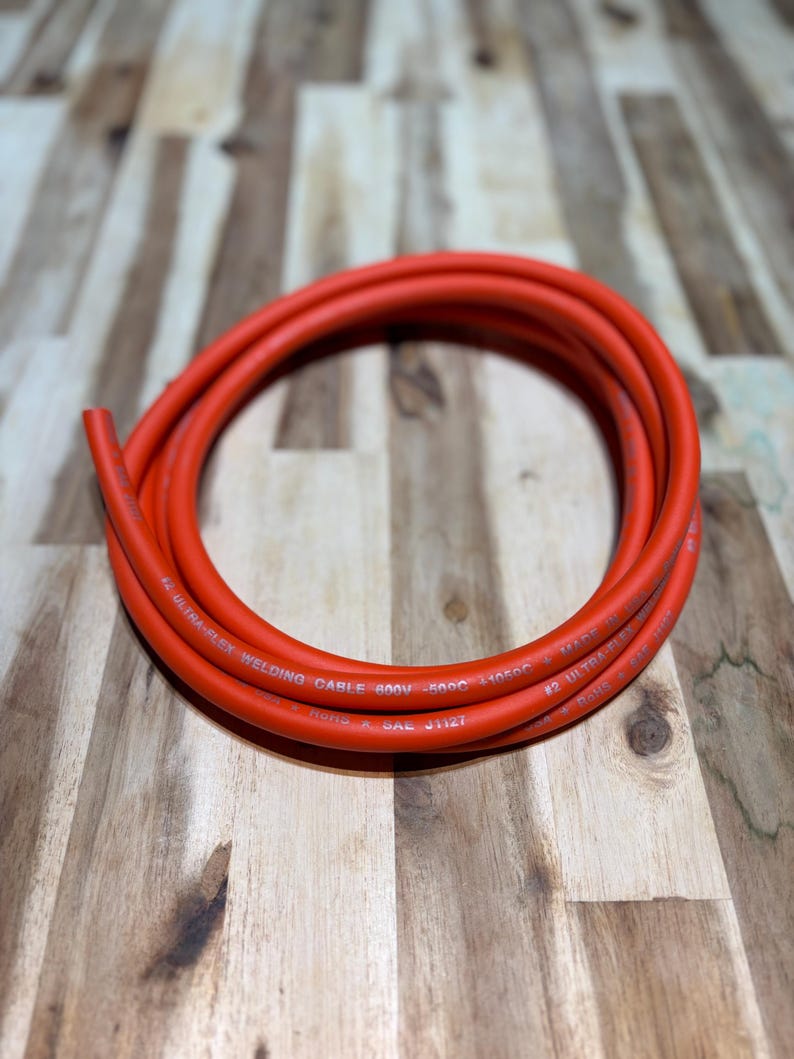May include: Coiled orange welding cable. The cable is labeled with text including "#2 ULTRA-FLEX WELDING CABLE 600V -50C 31050C". The cable is made in Hong Kong and meets SAE J1127 standards.