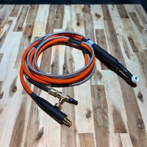May include: A coiled orange and gray welding torch cable with a black handle and nozzle. The cable has a black connector and a brass fitting. The background is a wooden surface.