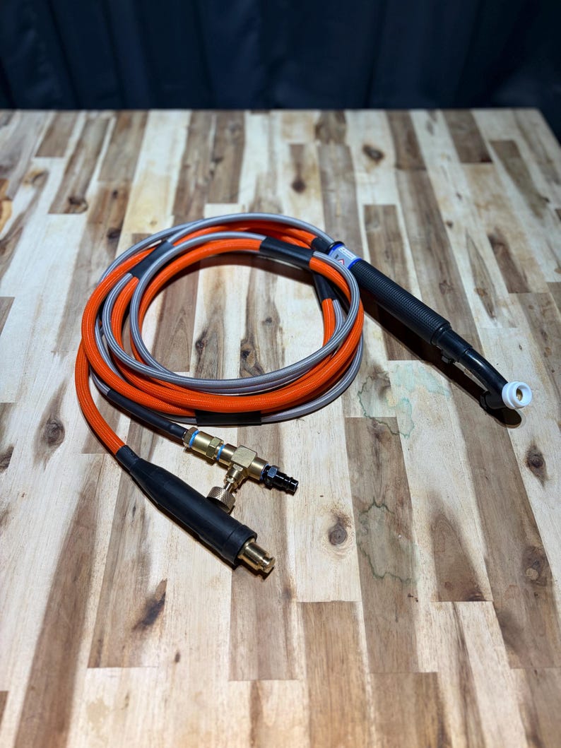 May include: A coiled orange and gray welding torch cable with a black handle and nozzle. The cable has a black connector and a brass fitting. The background is a wooden surface.