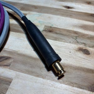 May include: Close-up of a black and gold electrical connector with a silver and purple braided cable. The connector has a black rubberized grip and a gold-colored metal tip. The cable is partially visible.
