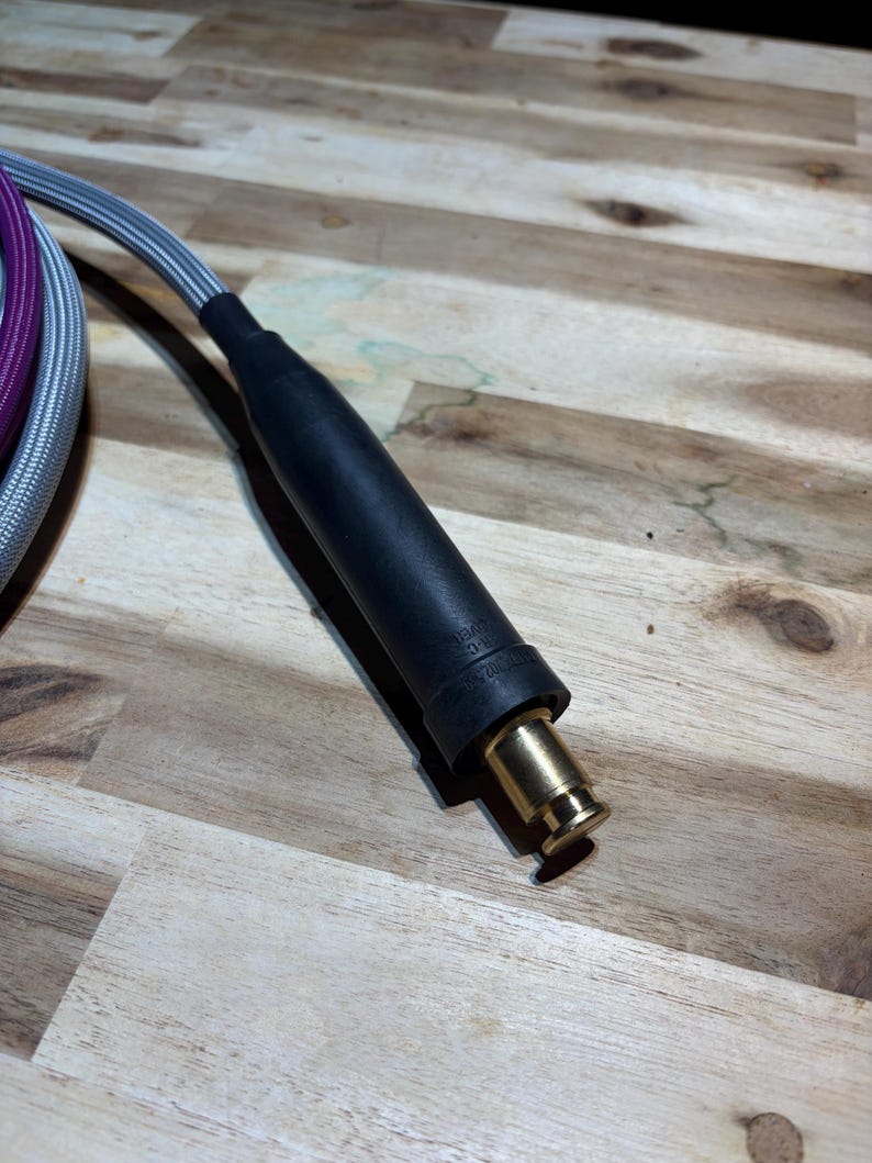 May include: Close-up of a black and gold electrical connector with a silver and purple braided cable. The connector has a black rubberized grip and a gold-colored metal tip. The cable is partially visible.