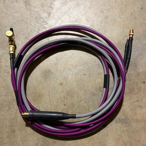 May include: Coiled audio cable with purple and gray braided sheathing. The cable has gold-colored connectors and a black connector. The cable is designed for audio equipment.