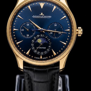 Könnte beinhalten: Eine goldfarbene Armbanduhr mit tiefblauem Zifferblatt, Mondphasenanzeige, zwei Hilfszifferblättern und goldenen Stundenmarkierungen. Die Uhr hat ein schwarzes Lederarmband mit strukturierter Oberfläche und zeigt den Schriftzug Jaeger-LeCoultre und Swiss Made.