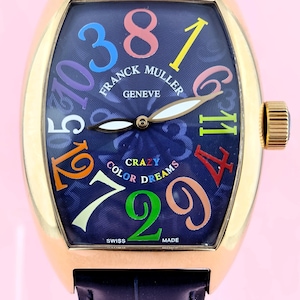 Könnte beinhalten: Eine goldfarbene Armbanduhr mit einem marineblauen Zifferblatt mit großen, farbigen Ziffern. Auf dem Zifferblatt stehen die Worte "CRAZY COLOR DREAMS" und "FRANCK MULLER GENEVE". Die Uhr hat ein marineblaues Lederarmband.