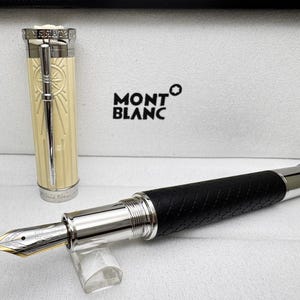 May include: A Montblanc fountain pen with a black textured barrel and silver detailing. The pen has a gold nib and a cream-coloured cap with a silver design. The Montblanc logo is visible in the background.
