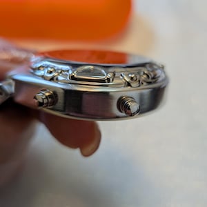 Pode incluir: Close-up de um rel&oacute;gio de pulso prateado com mostrador vermelho. O rel&oacute;gio tem dois bot&otilde;es na lateral e detalhes decorativos ao redor do mostrador. O rel&oacute;gio &eacute; segurado por uma pessoa.