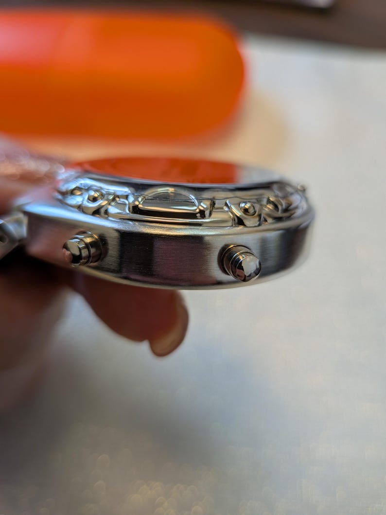Pode incluir: Close-up de um rel&oacute;gio de pulso prateado com mostrador vermelho. O rel&oacute;gio tem dois bot&otilde;es na lateral e detalhes decorativos ao redor do mostrador. O rel&oacute;gio &eacute; segurado por uma pessoa.