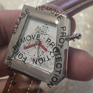 May include: A silver wristwatch with a white face featuring a musical instrument design and red hands. The watch has a brown leather band and is covered with a protective film that reads "PLEASE REMOVE PROTECTION".