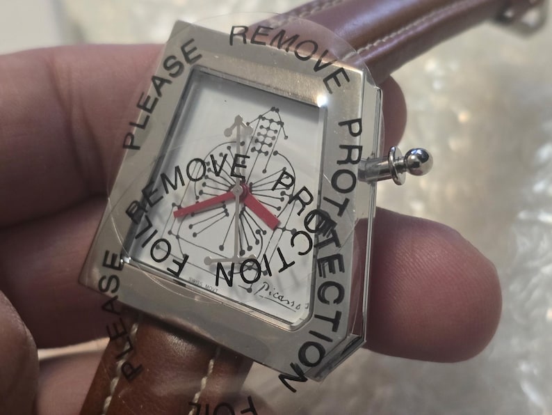 May include: A silver wristwatch with a white face featuring a musical instrument design and red hands. The watch has a brown leather band and is covered with a protective film that reads "PLEASE REMOVE PROTECTION".