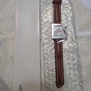 May include: A brown leather watch with a square silver face and a brown leather band. The watch face has a compass design and the words "REMOVE PROTECTION FOIL" are printed around the edge. The watch is packaged in bubble wrap and a white box.