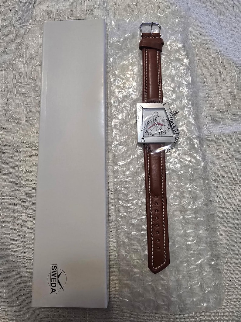 May include: A brown leather watch with a square silver face and a brown leather band. The watch face has a compass design and the words "REMOVE PROTECTION FOIL" are printed around the edge. The watch is packaged in bubble wrap and a white box.