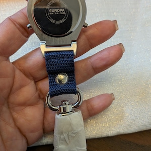 Pode incluir: Um rel&oacute;gio prateado com uma pulseira azul marinho e um fecho de metal. O mostrador do rel&oacute;gio n&atilde;o &eacute; vis&iacute;vel, mas a parte de tr&aacute;s do rel&oacute;gio &eacute; vis&iacute;vel, com o texto "EUROPA WATCH.COM" e "CHINA MOVEMENT". O rel&oacute;gio est&aacute; preso a um porta-chaves.