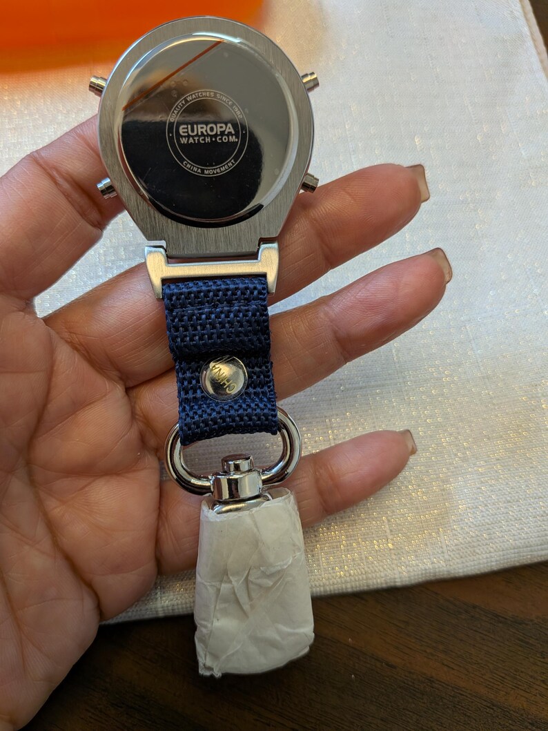 Pode incluir: Um rel&oacute;gio prateado com uma pulseira azul marinho e um fecho de metal. O mostrador do rel&oacute;gio n&atilde;o &eacute; vis&iacute;vel, mas a parte de tr&aacute;s do rel&oacute;gio &eacute; vis&iacute;vel, com o texto "EUROPA WATCH.COM" e "CHINA MOVEMENT". O rel&oacute;gio est&aacute; preso a um porta-chaves.