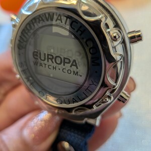 Pode incluir: Um rel&oacute;gio digital Europa Watch prateado com uma pulseira azul marinho. O mostrador do rel&oacute;gio exibe o logotipo Europa Watch e o texto "SEAL OF QUALITY". O rel&oacute;gio tem tr&ecirc;s bot&otilde;es laterais.
