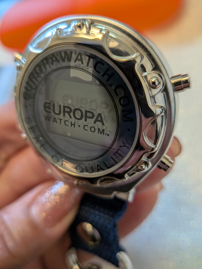 Pode incluir: Um rel&oacute;gio digital Europa Watch prateado com uma pulseira azul marinho. O mostrador do rel&oacute;gio exibe o logotipo Europa Watch e o texto "SEAL OF QUALITY". O rel&oacute;gio tem tr&ecirc;s bot&otilde;es laterais.