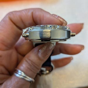 Pode incluir: Um rel&oacute;gio de pulso prateado com uma pulseira preta. O mostrador do rel&oacute;gio &eacute; adornado com pequenos enfeites brilhantes. O rel&oacute;gio &eacute; segurado em uma m&atilde;o com um anel de prata.