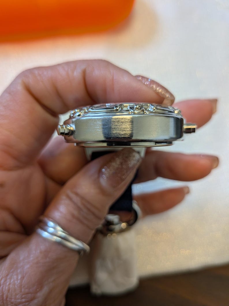 Pode incluir: Um rel&oacute;gio de pulso prateado com uma pulseira preta. O mostrador do rel&oacute;gio &eacute; adornado com pequenos enfeites brilhantes. O rel&oacute;gio &eacute; segurado em uma m&atilde;o com um anel de prata.