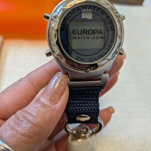 Pode incluir: Um rel&oacute;gio Europa Watch-Com prateado com uma pulseira azul marinho. O mostrador do rel&oacute;gio exibe o logotipo Europa Watch-Com e as palavras "Seal of Quality". O rel&oacute;gio &eacute; segurado por uma pessoa.