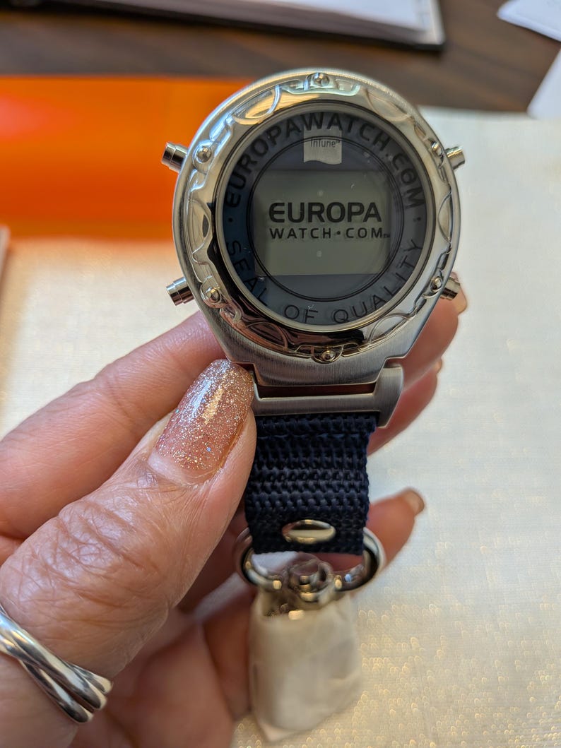 Pode incluir: Um rel&oacute;gio Europa Watch-Com prateado com uma pulseira azul marinho. O mostrador do rel&oacute;gio exibe o logotipo Europa Watch-Com e as palavras "Seal of Quality". O rel&oacute;gio &eacute; segurado por uma pessoa.
