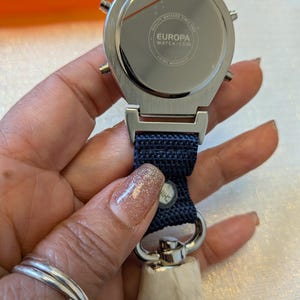 Pode incluir: Um rel&oacute;gio Europa prateado com uma pulseira de tecido azul marinho. O rel&oacute;gio tem um mostrador redondo com tr&ecirc;s pequenos bot&otilde;es na lateral. A parte de tr&aacute;s do rel&oacute;gio tem o texto "EUROPA WATCH.COM" e "CHINA MOVEMENT".