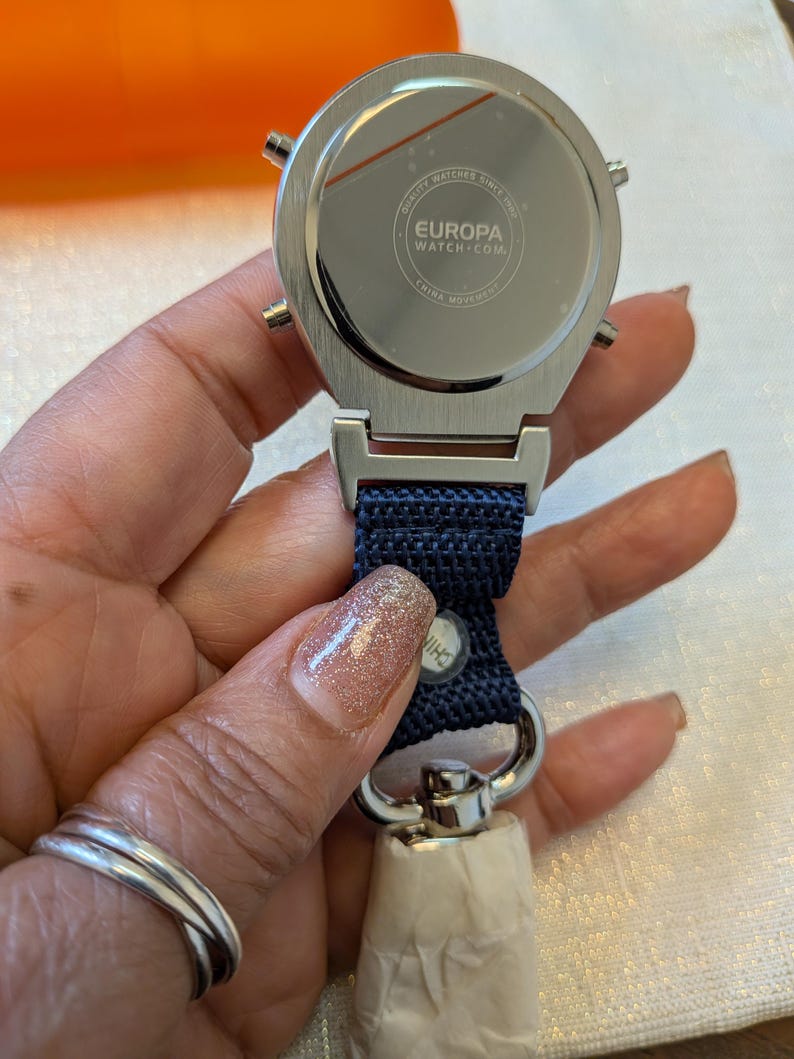 Pode incluir: Um rel&oacute;gio Europa prateado com uma pulseira de tecido azul marinho. O rel&oacute;gio tem um mostrador redondo com tr&ecirc;s pequenos bot&otilde;es na lateral. A parte de tr&aacute;s do rel&oacute;gio tem o texto "EUROPA WATCH.COM" e "CHINA MOVEMENT".