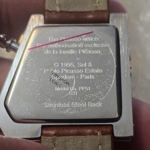May include: Close-up of the back of a stainless steel watch with a brown leather strap. The watch face has text that reads "The Picasso watch" and "Stainless Steel Back". The watch is held by a person.