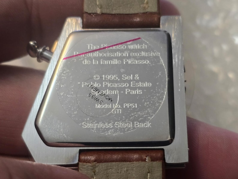 May include: Close-up of the back of a stainless steel watch with a brown leather strap. The watch face has text that reads "The Picasso watch" and "Stainless Steel Back". The watch is held by a person.