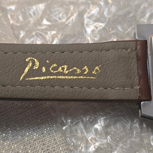 May include: Close-up of a wristwatch with a gray leather band. The word "Picasso" is printed in gold script on the band. The watch face is silver with a brown leather strap.