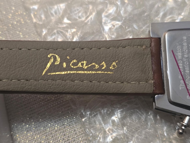 May include: Close-up of a wristwatch with a gray leather band. The word "Picasso" is printed in gold script on the band. The watch face is silver with a brown leather strap.
