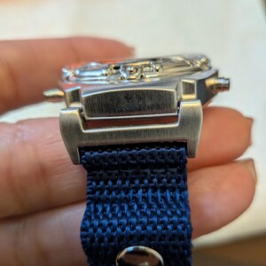 Pode incluir: Um rel&oacute;gio de pulso prateado com uma pulseira de tecido azul escuro. O mostrador do rel&oacute;gio &eacute; vis&iacute;vel e a pulseira &eacute; presa com um fecho de metal. O rel&oacute;gio tem um design cl&aacute;ssico e elegante.
