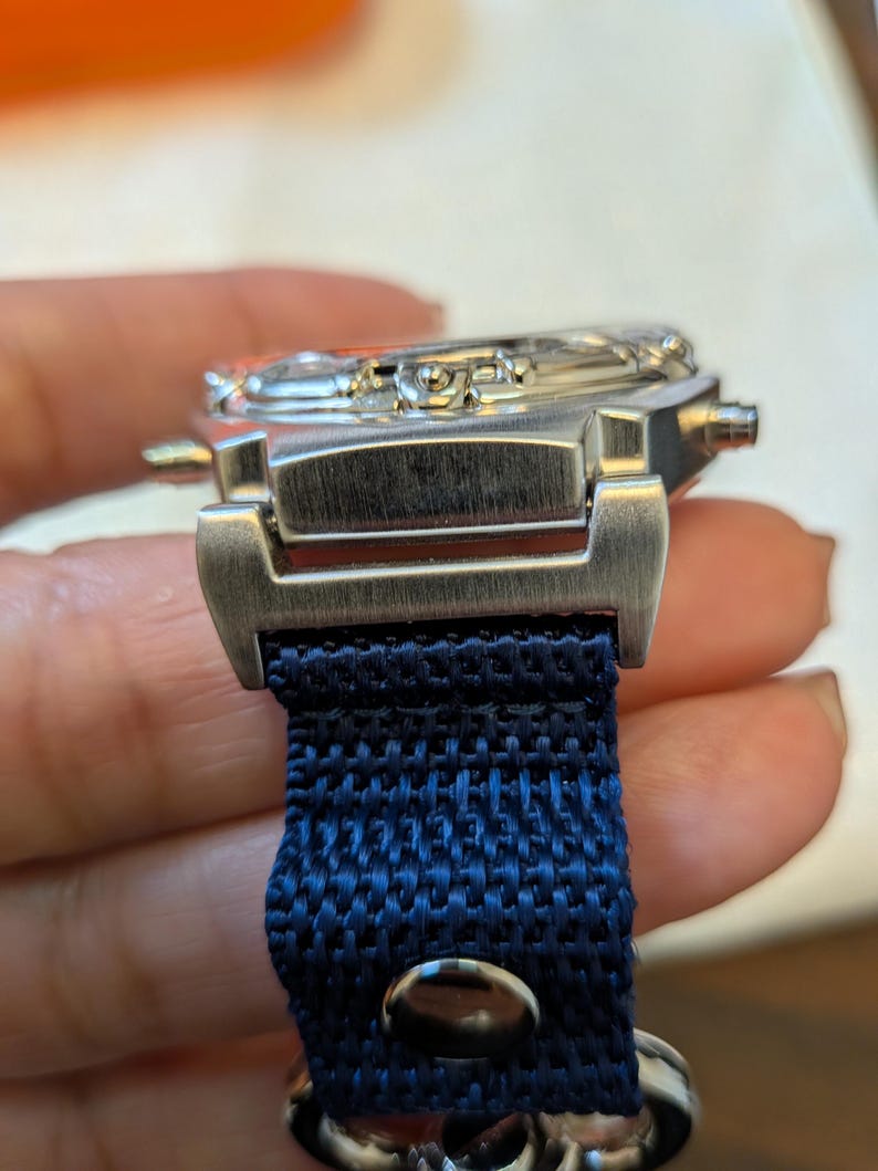 Pode incluir: Um rel&oacute;gio de pulso prateado com uma pulseira de tecido azul escuro. O mostrador do rel&oacute;gio &eacute; vis&iacute;vel e a pulseira &eacute; presa com um fecho de metal. O rel&oacute;gio tem um design cl&aacute;ssico e elegante.