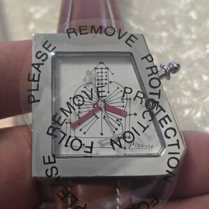 May include: A silver-toned wristwatch with a geometric face design and red hands. The watch has a brown leather band and is covered with a protective film that reads "PLEASE REMOVE PROTECTION". The watch face is marked "Picasso".