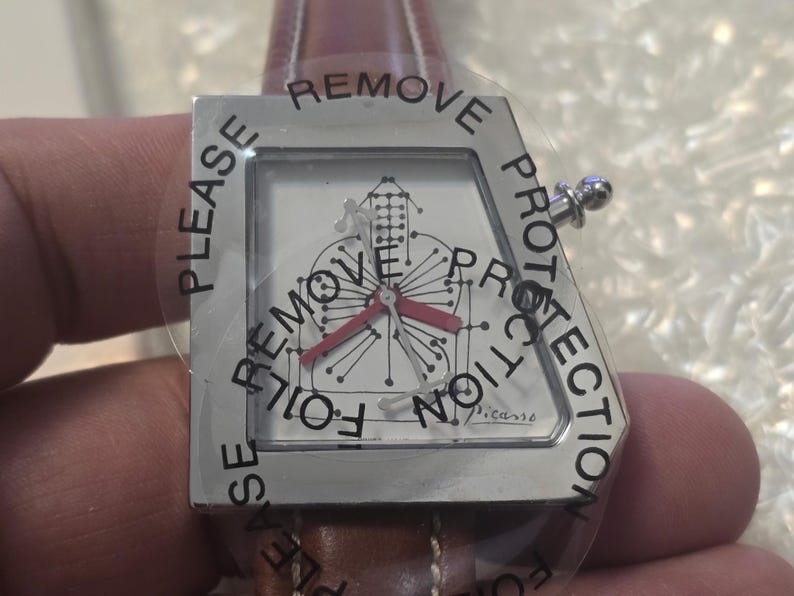 May include: A silver-toned wristwatch with a geometric face design and red hands. The watch has a brown leather band and is covered with a protective film that reads "PLEASE REMOVE PROTECTION". The watch face is marked "Picasso".