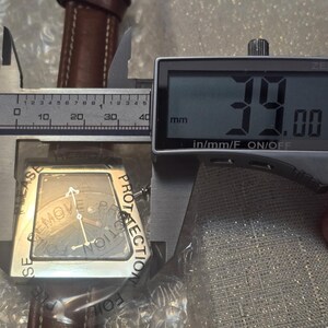 May include: A silver-toned watch with a brown leather band is measured with a digital caliper. The watch face is square with a dark background and silver hands. The caliper displays a measurement of 39.00 mm.