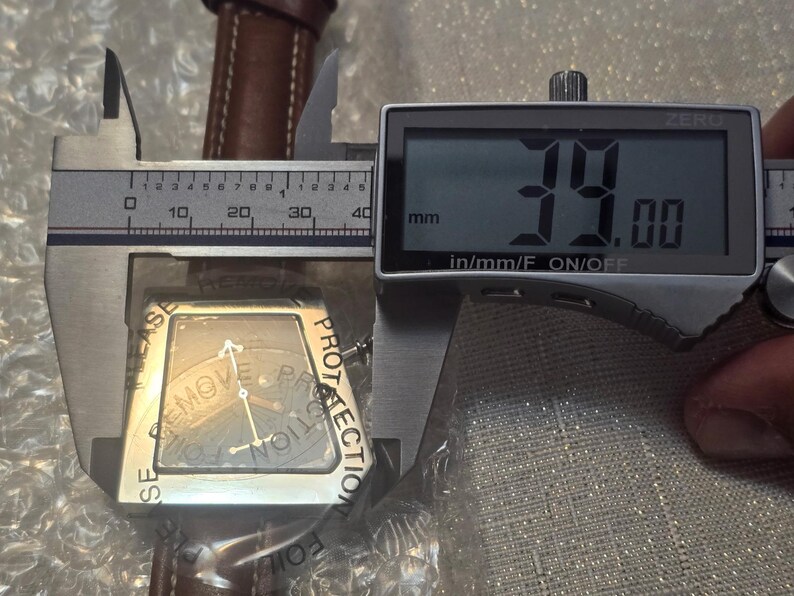 May include: A silver-toned watch with a brown leather band is measured with a digital caliper. The watch face is square with a dark background and silver hands. The caliper displays a measurement of 39.00 mm.