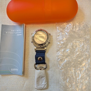 Pode incluir: Um rel&oacute;gio Europa Watch Co. prateado com uma pulseira azul marinho e uma etiqueta branca. O rel&oacute;gio est&aacute; ao lado de um livreto, um saco pl&aacute;stico transparente e um recipiente laranja em forma de c&aacute;psula.