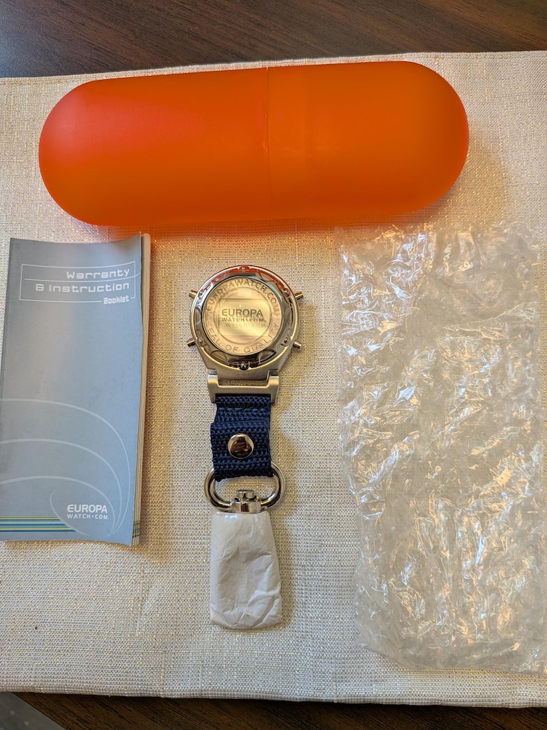 Pode incluir: Um rel&oacute;gio Europa Watch Co. prateado com uma pulseira azul marinho e uma etiqueta branca. O rel&oacute;gio est&aacute; ao lado de um livreto, um saco pl&aacute;stico transparente e um recipiente laranja em forma de c&aacute;psula.