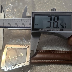 May include: A digital caliper measuring a watch component. The digital display reads 38.50 mm. The watch component is silver-colored with a protective film that reads "PLEASE REMOVE PROTECTION". The watch band is brown leather.