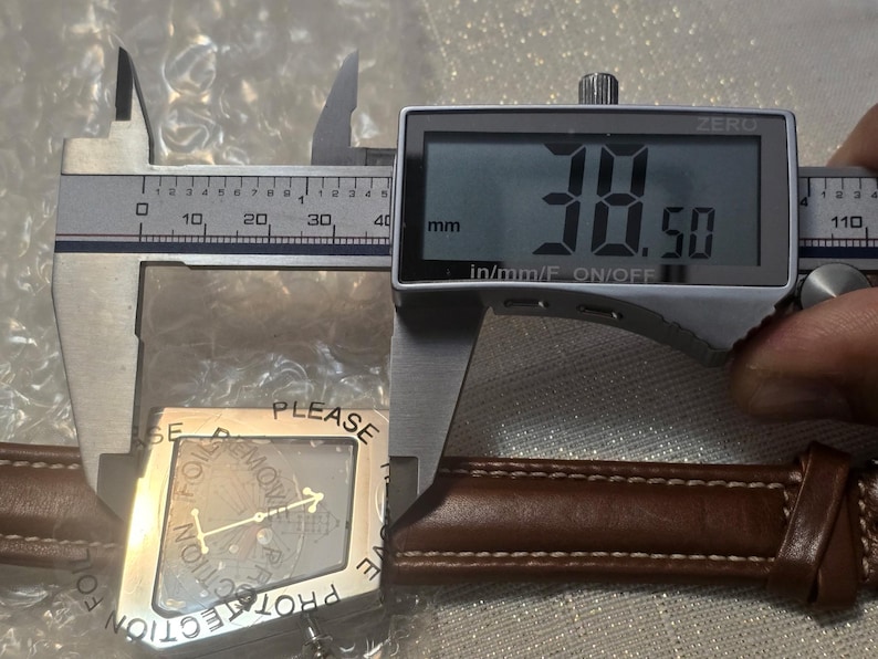 May include: A digital caliper measuring a watch component. The digital display reads 38.50 mm. The watch component is silver-colored with a protective film that reads "PLEASE REMOVE PROTECTION". The watch band is brown leather.