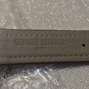 May include: Close-up of a brown leather watch strap. The underside is a light beige color with the words "GENUINE LEATHER" and "CHINA" stamped into the material. The strap is stitched with a light beige thread.