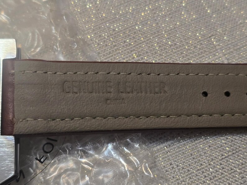 May include: Close-up of a brown leather watch strap. The underside is a light beige color with the words "GENUINE LEATHER" and "CHINA" stamped into the material. The strap is stitched with a light beige thread.