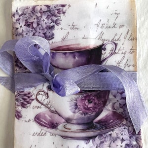 May include: A vintage teacup and saucer with purple floral designs, tied with a lavender ribbon and surrounded by purple flowers. The background is a vintage paper with handwritten text.